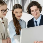Three young business people, 2 women and a man, stand around a single laptop looking at the screen.